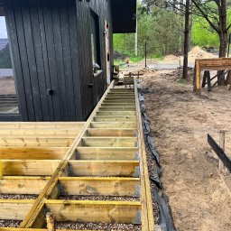 Michał Wieczorek DOMY DREWNIANE ,SZKIELETOWE. OBSŁUGA KOMPLEKSOWA - Konstrukcja drewnianego tarasu w trakcie budowy, widoczne legary na podsypce z kamienia, przy ciemnej elewacji domu z oknami.