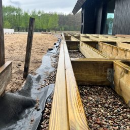 Michał Wieczorek DOMY DREWNIANE ,SZKIELETOWE. OBSŁUGA KOMPLEKSOWA - Konstrukcja drewnianego tarasu w trakcie budowy, widoczne legary, podłoże z kamyków, geowłóknina i czarny budynek w tle.