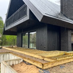 Michał Wieczorek DOMY DREWNIANE ,SZKIELETOWE. OBSŁUGA KOMPLEKSOWA - Nowoczesny dom z czarną elewacją i kominem z cegły, w trakcie budowy tarasu z drewnianą konstrukcją na betonowych fundamentach, widok na drzewa w tle, pochmurna pogoda.