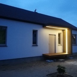 Michał Wieczorek DOMY DREWNIANE ,SZKIELETOWE. OBSŁUGA KOMPLEKSOWA - Wejście do białego domu z drewnianym podestem i oświetleniem LED wzdłuż ścianki, widoczne wczesnym wieczorem.