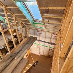 Michał Wieczorek DOMY DREWNIANE ,SZKIELETOWE. OBSŁUGA KOMPLEKSOWA - Wnętrze w trakcie budowy domu szkieletowego: widoczne drewniane ramy konstrukcyjne, izolacja termiczna z folią aluminiową na poddaszu, okno dachowe z zieloną taśmą uszczelniającą, fragment schodów...