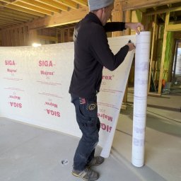 Michał Wieczorek DOMY DREWNIANE ,SZKIELETOWE. OBSŁUGA KOMPLEKSOWA - Dwóch monterów rozkłada membranę izolacyjną Siga Majrex 200 wewnątrz surowego domu szkieletowego, widoczne drewniane ramy konstrukcyjne i ocieplenie między nimi.