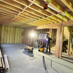 Michał Wieczorek DOMY DREWNIANE ,SZKIELETOWE. OBSŁUGA KOMPLEKSOWA - Wnętrze domu w trakcie budowy w technologii szkieletowej, widoczne ocieplone ściany wełną mineralną, drewniana konstrukcja nośna, pracownik z odkurzaczem przemysłowym oraz nagrzewnica, na podłodze...