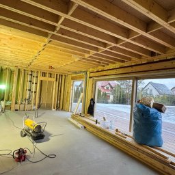 Michał Wieczorek DOMY DREWNIANE ,SZKIELETOWE. OBSŁUGA KOMPLEKSOWA - Wnętrze domu w trakcie budowy szkieletowej, widoczne drewniane elementy konstrukcyjne, oświetlenie budowlane, nagrzewnica, odkurzacz przemysłowy oraz okno z widokiem na zaśnieżony taras i zabudowę...