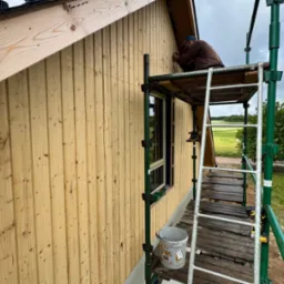 Prace wykończeniowe na elewacji domu z drewnianą boazerią. Mężczyzna na rusztowaniu przy oknie. Widok na pola i las w tle. Jasne drewno, naturalne światło dzienne.