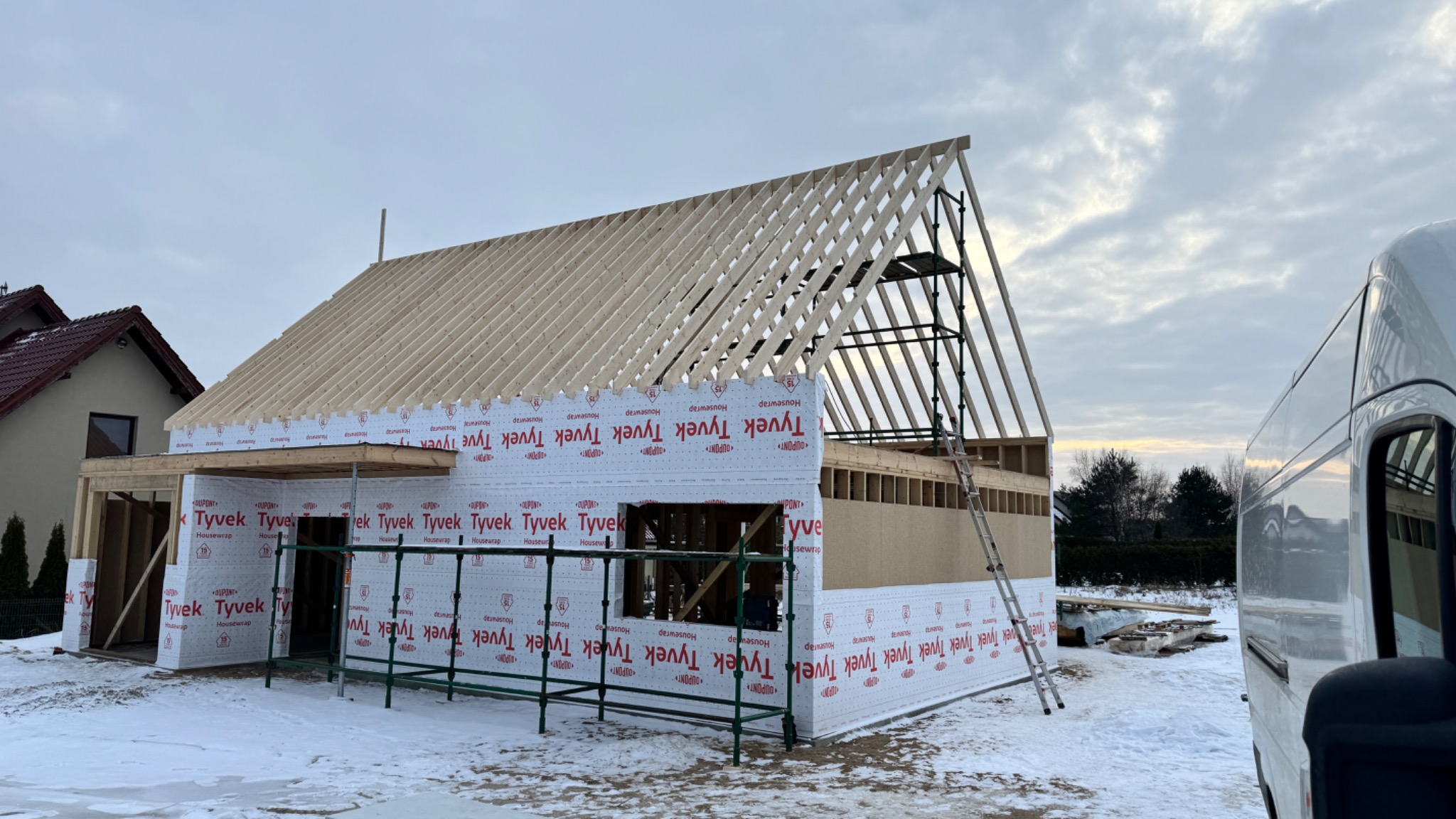 Budowa domu szkieletowego w zimowej scenerii: konstrukcja z drewnianych belek, pokryta membraną Tyvek, rusztowanie i drabina oparta o ścianę, widoczny fragment białego vana.