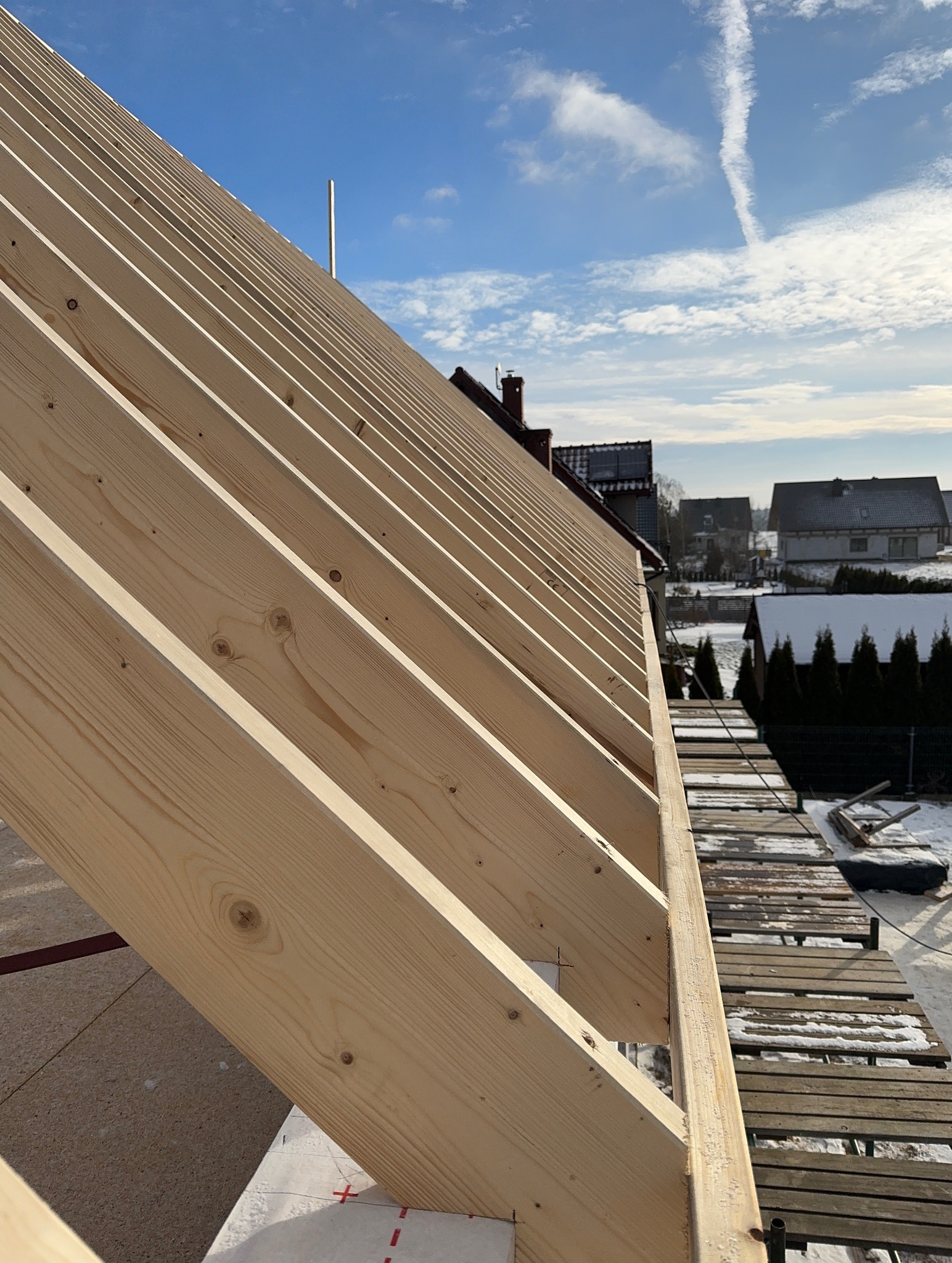 Konstrukcja drewniana dachu w trakcie budowy, widoczne elementy więźby dachowej na tle błękitnego nieba z chmurami i zabudowań w oddali.