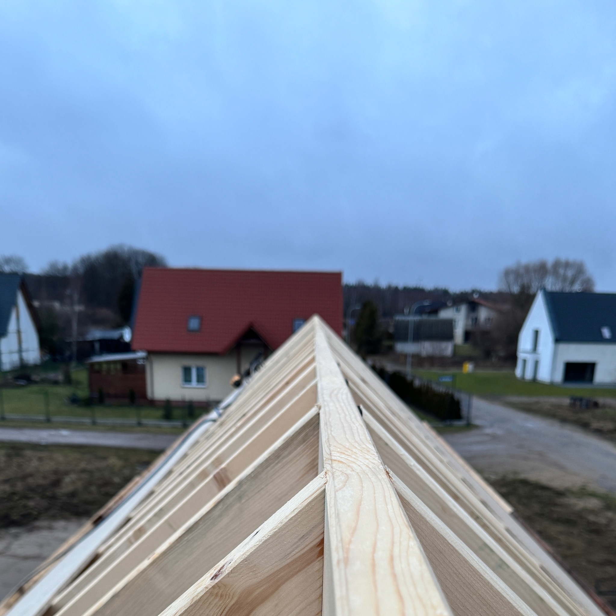 Konstrukcja dachu z drewnianych belek, widok z góry na tle zabudowy jednorodzinnej w pochmurny dzień.