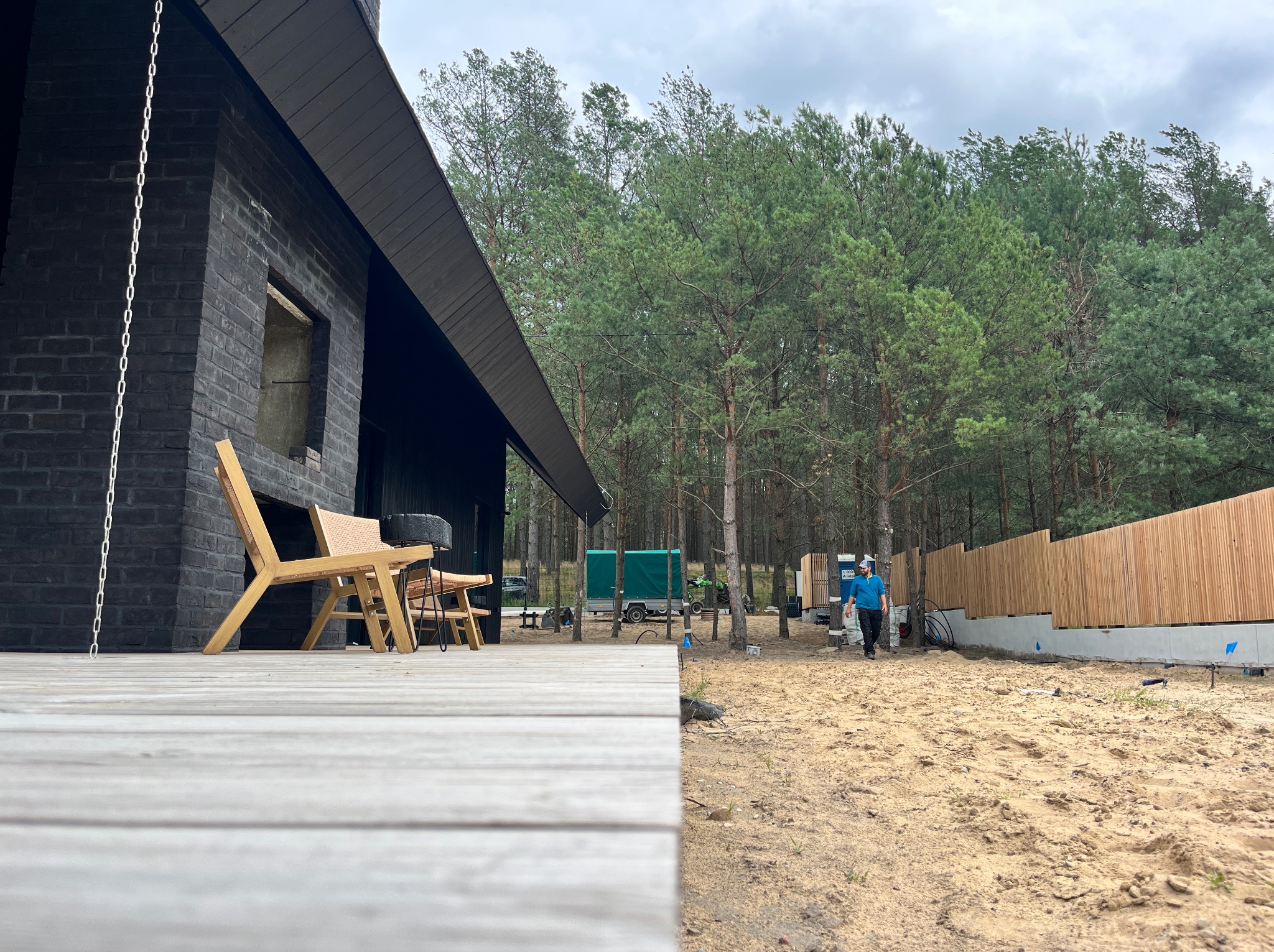 Ciemna elewacja domu w stylu nowoczesnej stodoły z drewnianym tarasem i meblami ogrodowymi, widok na teren budowy z ogrodzeniem i pracownikiem w tle.