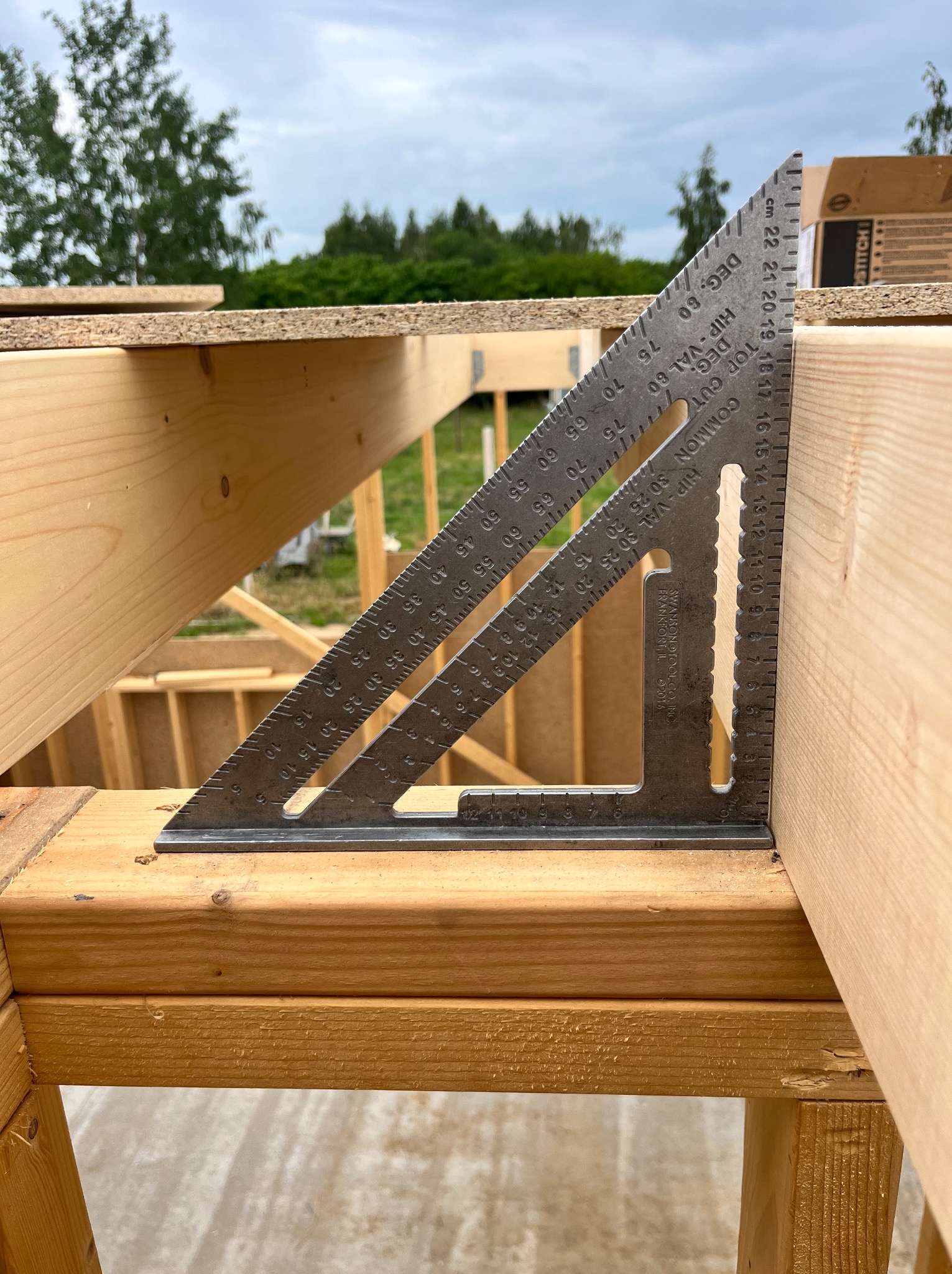 Metalowy kątownik stolarski na drewnianej konstrukcji szkieletowej domu w trakcie budowy, z widocznymi oznaczeniami kątów i miar.