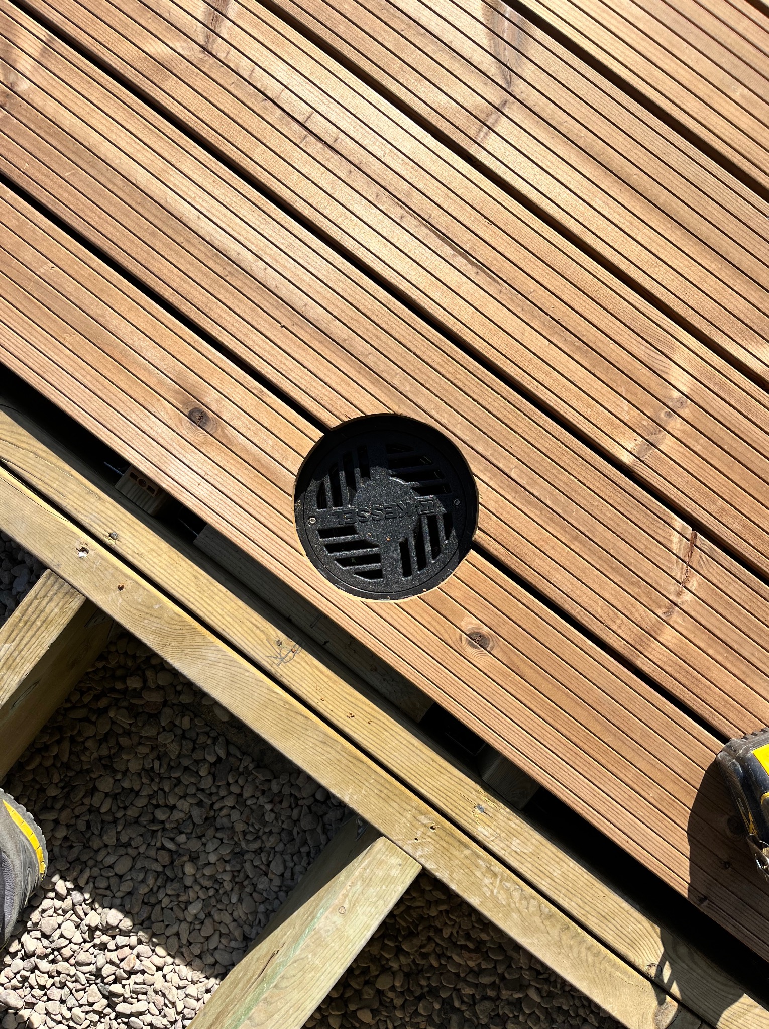 Drewniany taras z odpływem liniowym, widoczne elementy konstrukcyjne i podłoże z kamyków, fragment buta roboczego w rogu.