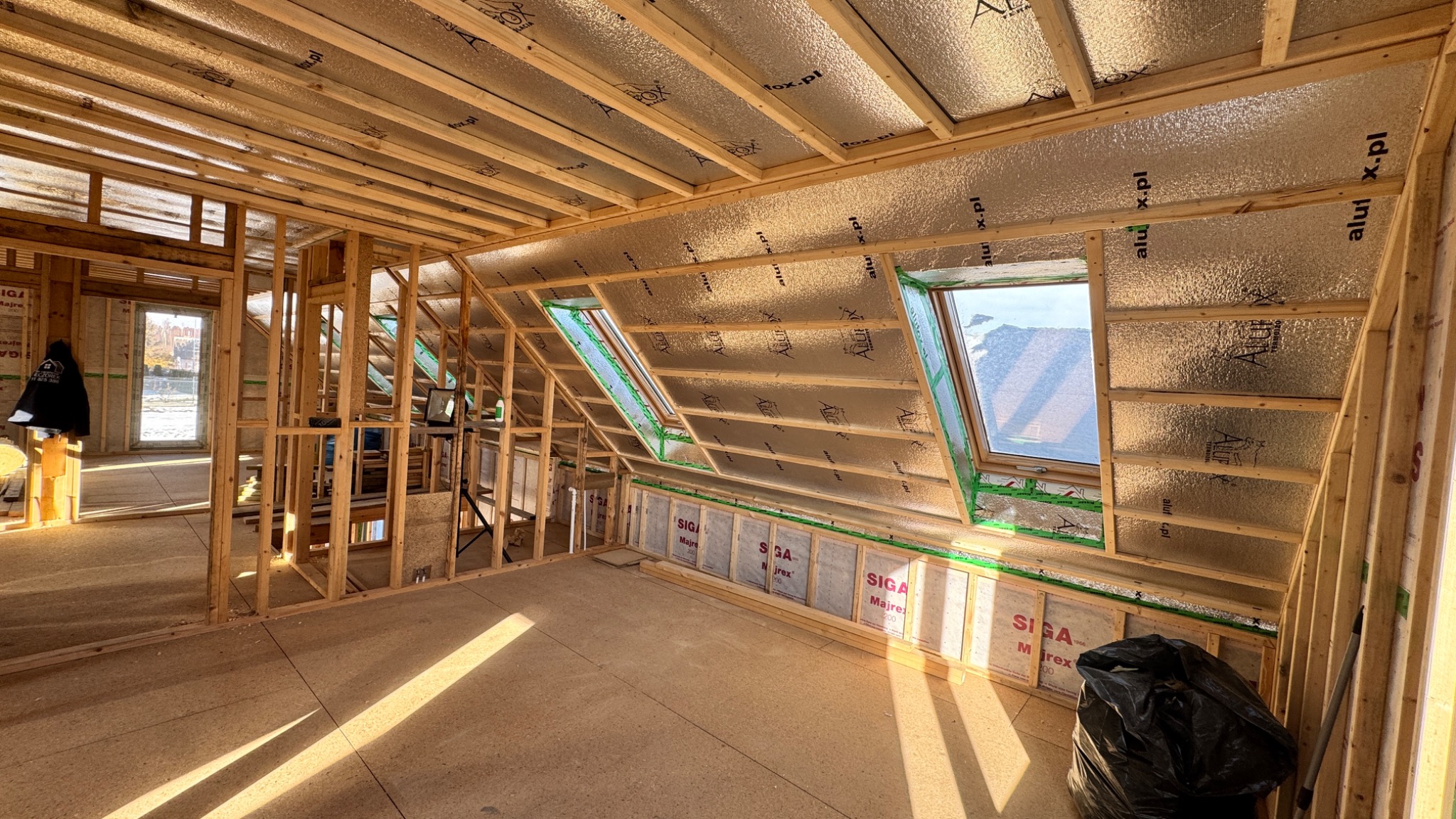 Poddasze w trakcie ocieplania, widoczne okna dachowe, folia aluminiowa izolacyjna na skosach, drewniana konstrukcja szkieletowa, częściowo ocieplona ściana z widocznymi membranami Siga Majrex...