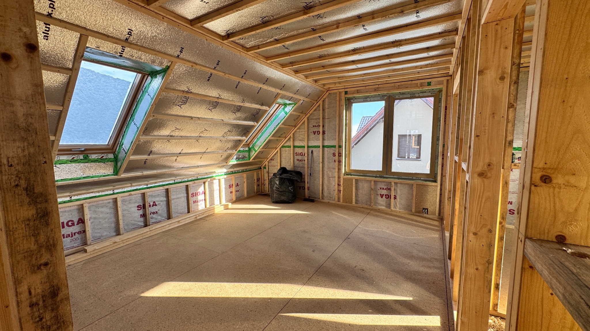 Poddasze w trakcie budowy, widoczne okna dachowe, izolacja folią aluminiową, drewniana konstrukcja szkieletowa ścian i stropu, oraz czarny worek na śmieci w rogu pomieszczenia.