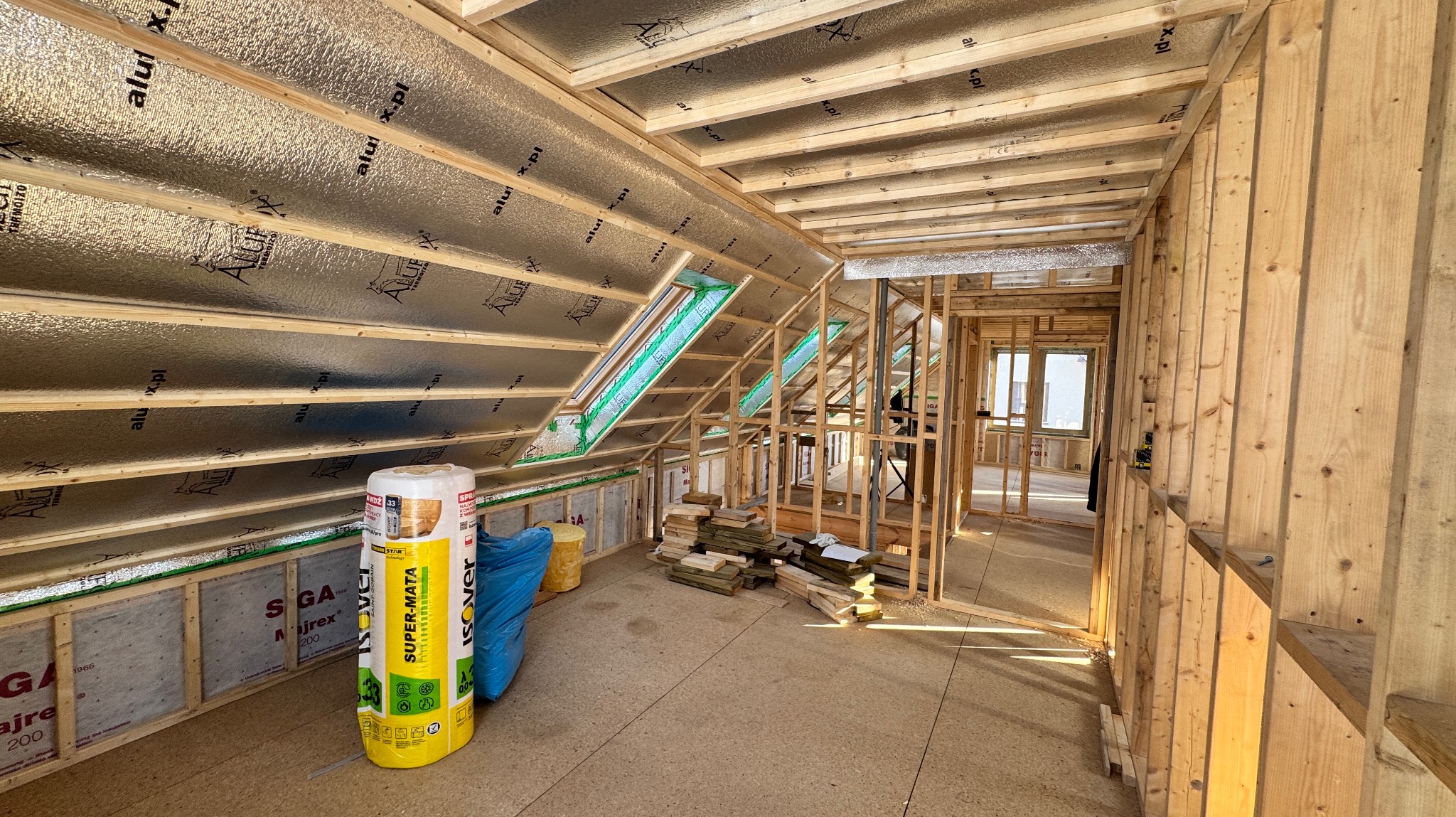 Wnętrze domu w trakcie budowy z widoczną konstrukcją szkieletową, izolacją termiczną na poddaszu i folią aluminiową, rolka wełny mineralnej Isover Super-Mata stoi na podłodze.