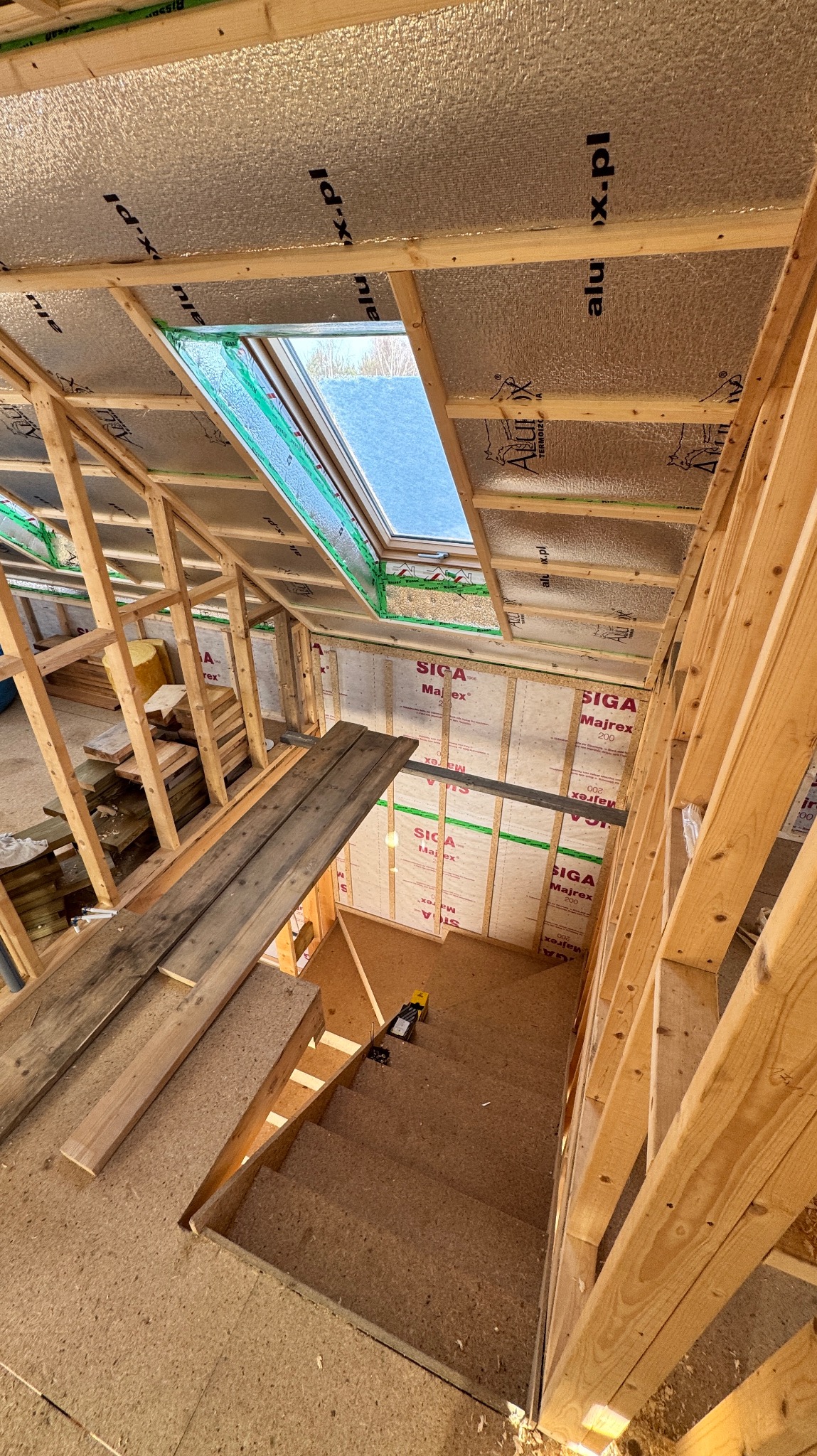 Wnętrze w trakcie budowy domu szkieletowego: widoczne drewniane ramy konstrukcyjne, izolacja termiczna z folią aluminiową na poddaszu, okno dachowe z zieloną taśmą uszczelniającą, fragment schodów...
