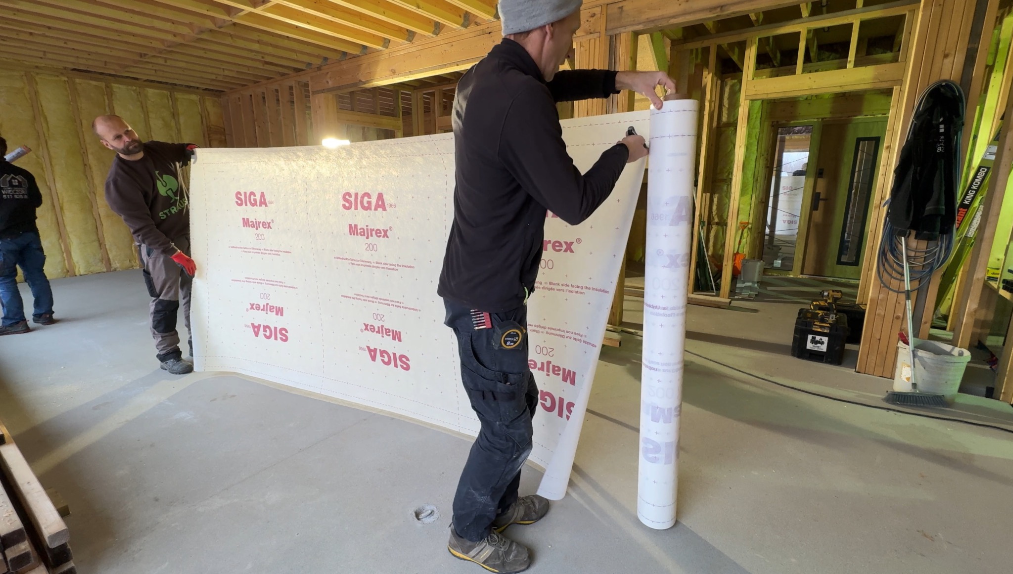 Dwóch monterów rozkłada membranę izolacyjną Siga Majrex 200 wewnątrz surowego domu szkieletowego, widoczne drewniane ramy konstrukcyjne i ocieplenie między nimi.