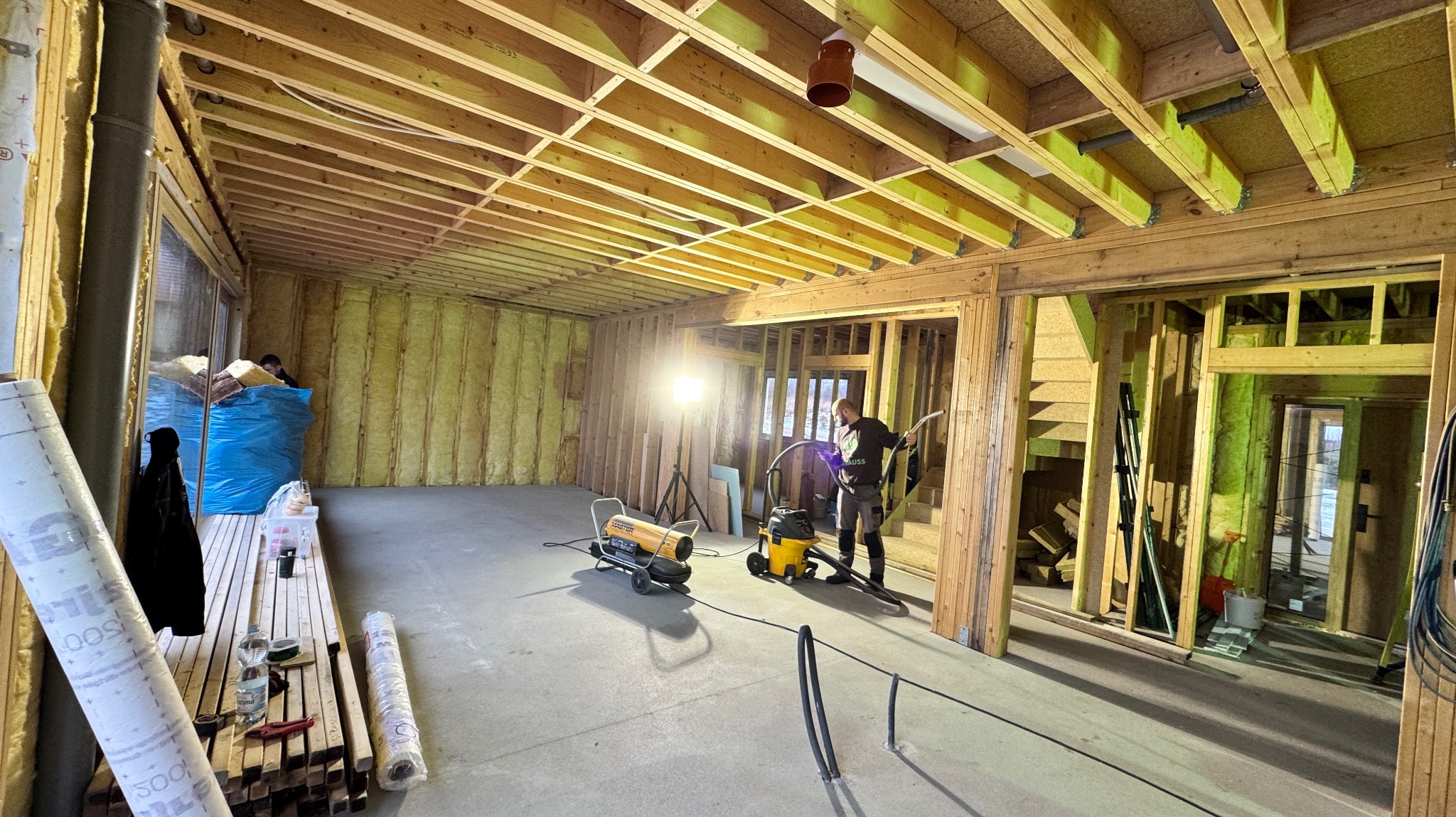 Wnętrze domu w trakcie budowy w technologii szkieletowej, widoczne ocieplone ściany wełną mineralną, drewniana konstrukcja nośna, pracownik z odkurzaczem przemysłowym oraz nagrzewnica, na podłodze...