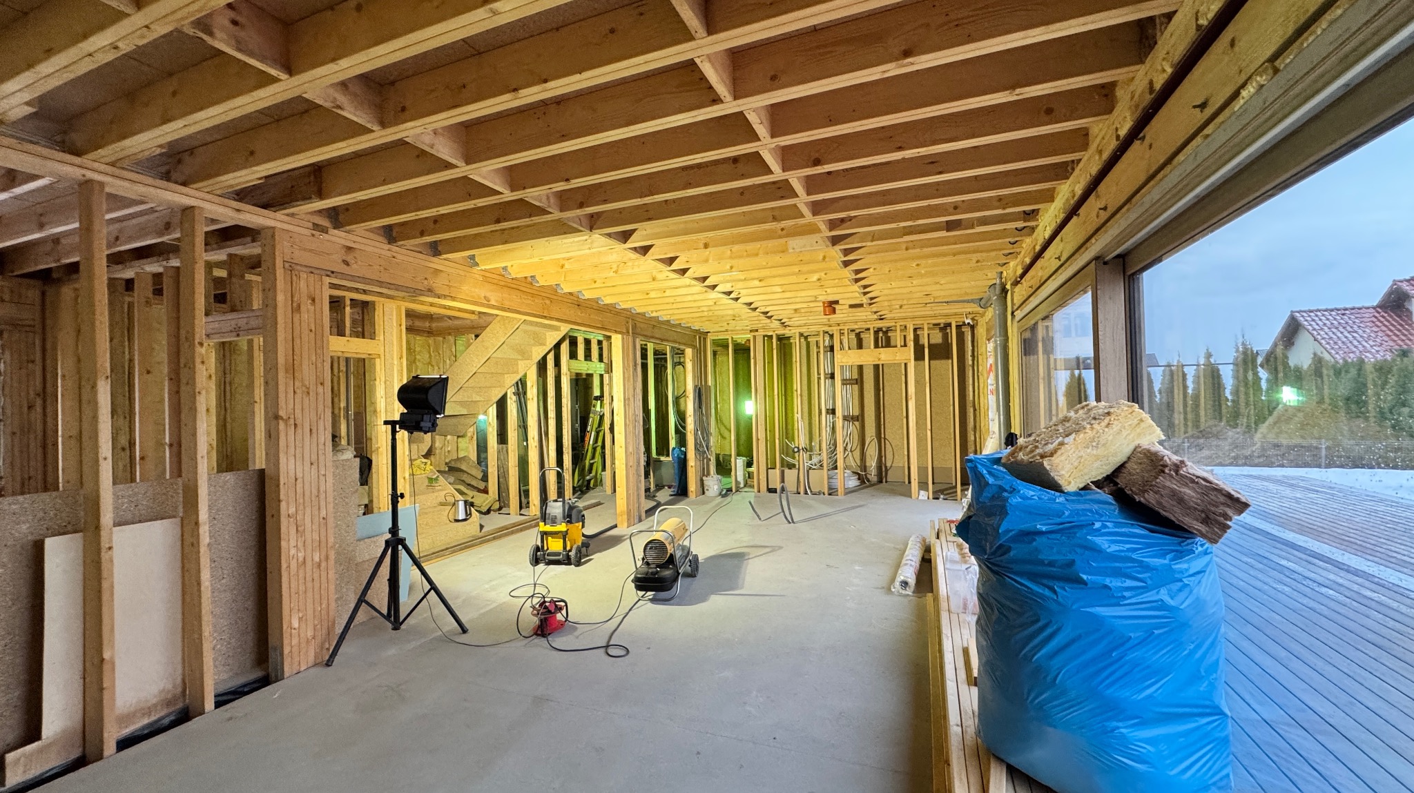 Wnętrze domu w trakcie budowy w technologii szkieletowej, widoczne drewniane ramy ścian, strop z belek, schody, oświetlenie robocze, izolacja w worku i widok na zewnątrz przez duże okno.
