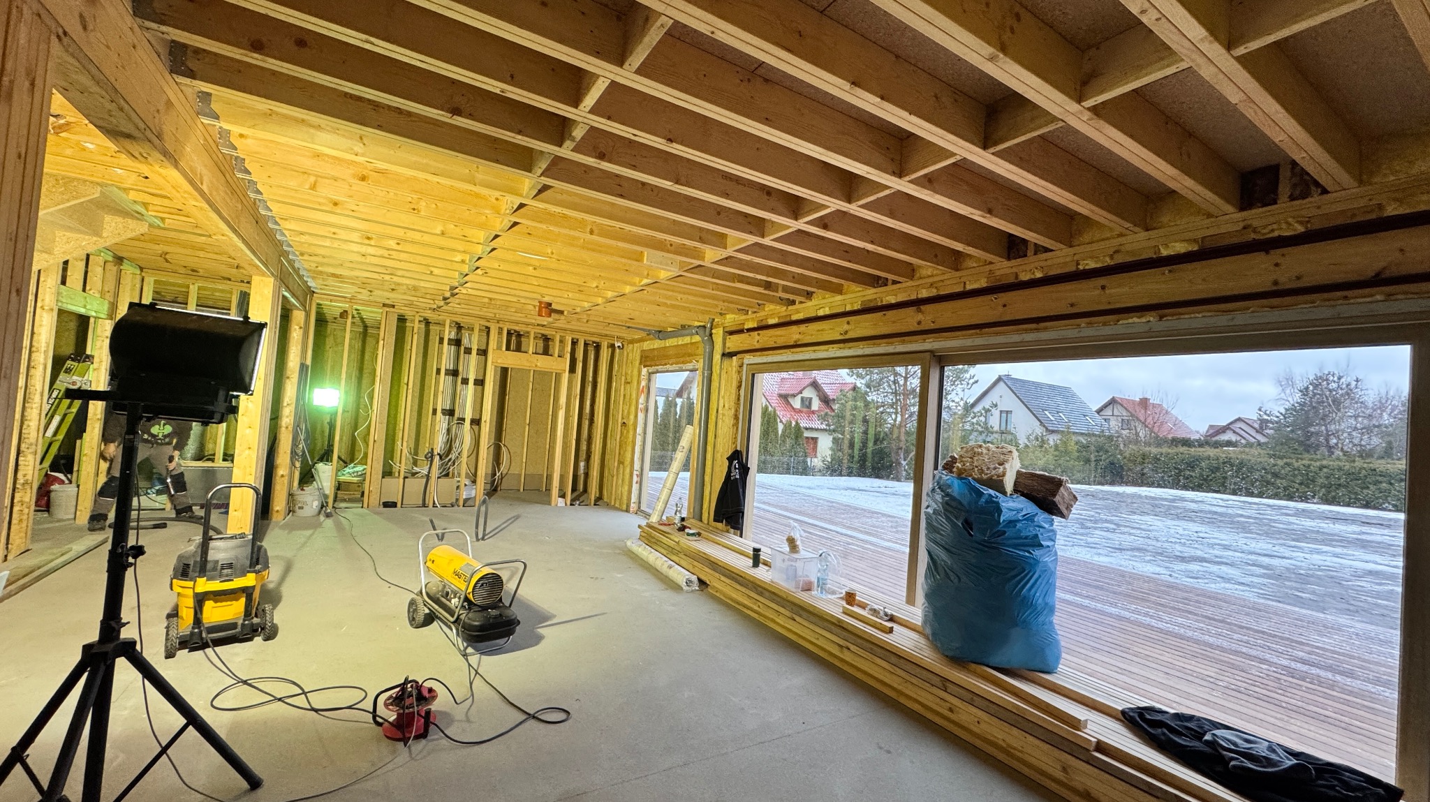 Wnętrze domu w trakcie budowy szkieletowej, widoczne drewniane elementy konstrukcyjne, oświetlenie budowlane, nagrzewnica, odkurzacz przemysłowy oraz okno z widokiem na zaśnieżony taras i zabudowę...