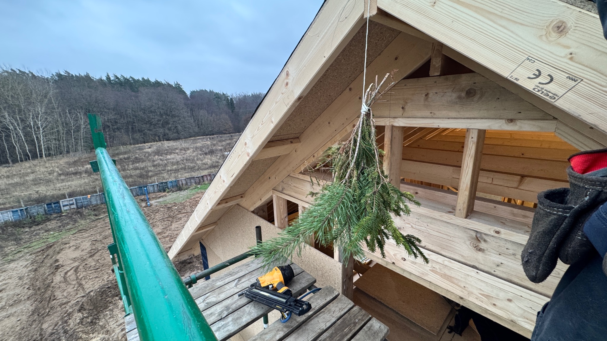 Wieniec zawieszony na drewnianej konstrukcji w trakcie budowy, widok z rusztowania, w tle zalesiony teren i pochmurne niebo.