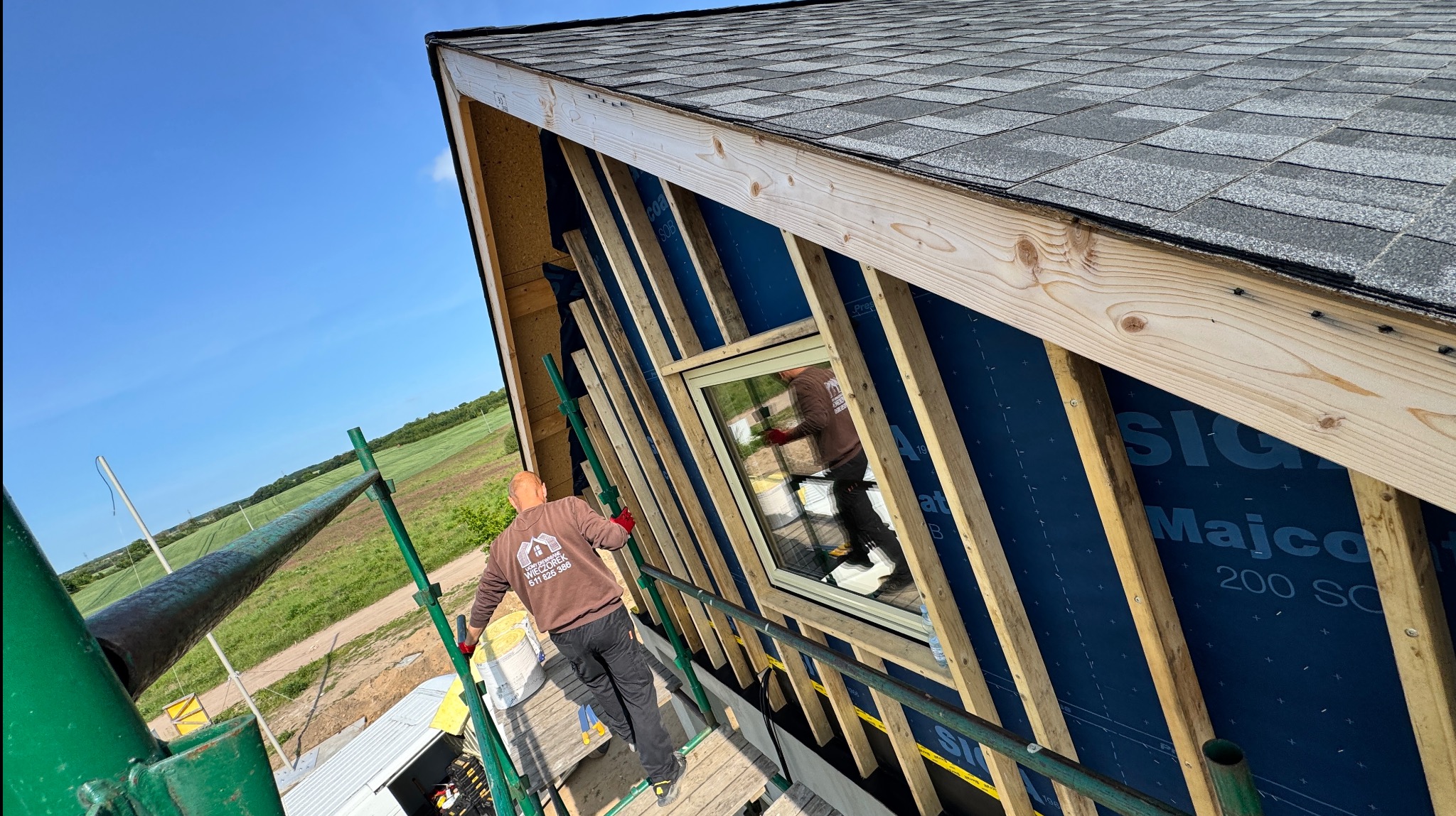 Montaż okna w konstrukcji dachu z widoczną membraną i łatami. Pracownik w brązowej bluzie z logo firmy na rusztowaniu. Widok z góry na plac budowy i zielone pola.