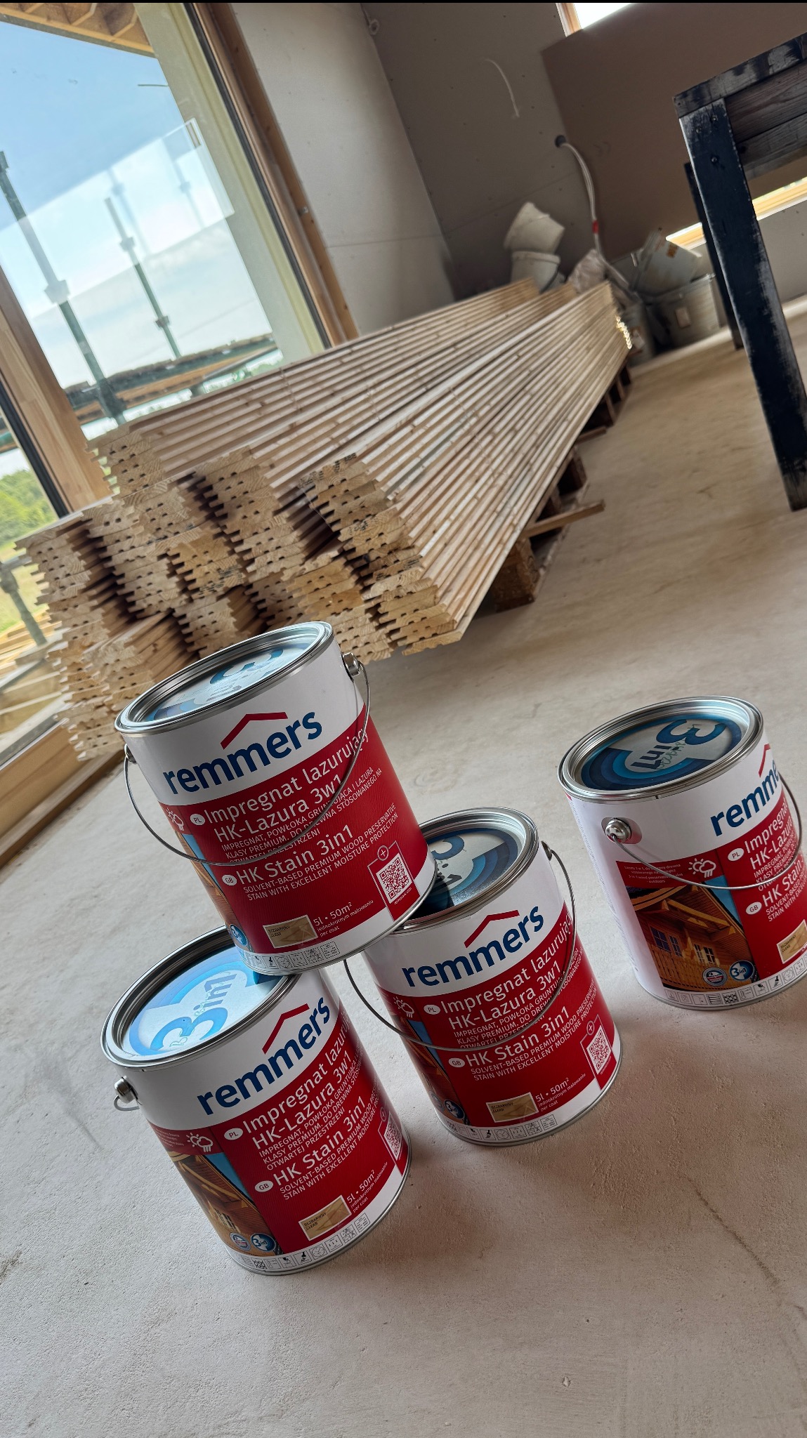 Cztery puszki z impregnatem Remmers HK-Lazura 3w1 na tle ułożonych desek i okna w surowym stanie. Widoczne elementy konstrukcji domu w trakcie budowy.