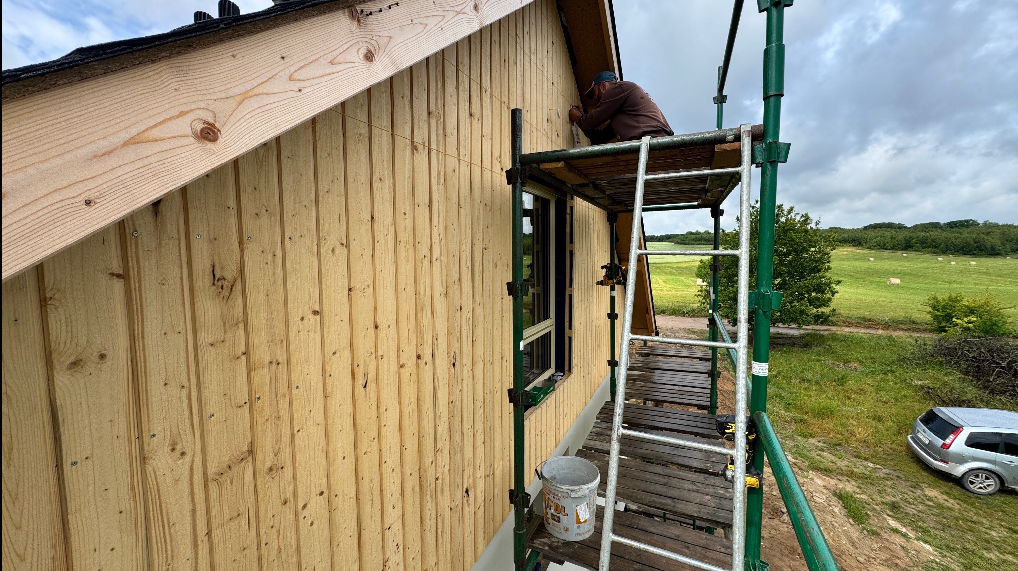 Prace wykończeniowe na elewacji domu z drewnianą boazerią. Mężczyzna na rusztowaniu przy oknie. Widok na pola i las w tle. Jasne drewno, naturalne światło dzienne.