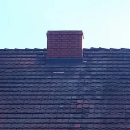 Czerwony ceglany komin wystający z ciemnego dachu pokrytego dachówką ceramiczną na tle jasnego nieba.