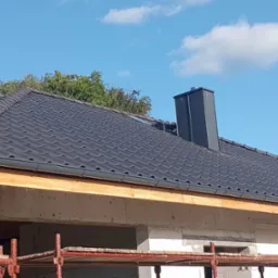Nowy dach z ciemnej dachówki ceramicznej i szarym kominem na tle błękitnego nieba z chmurami, widoczne rusztowanie i surowy stan budynku.