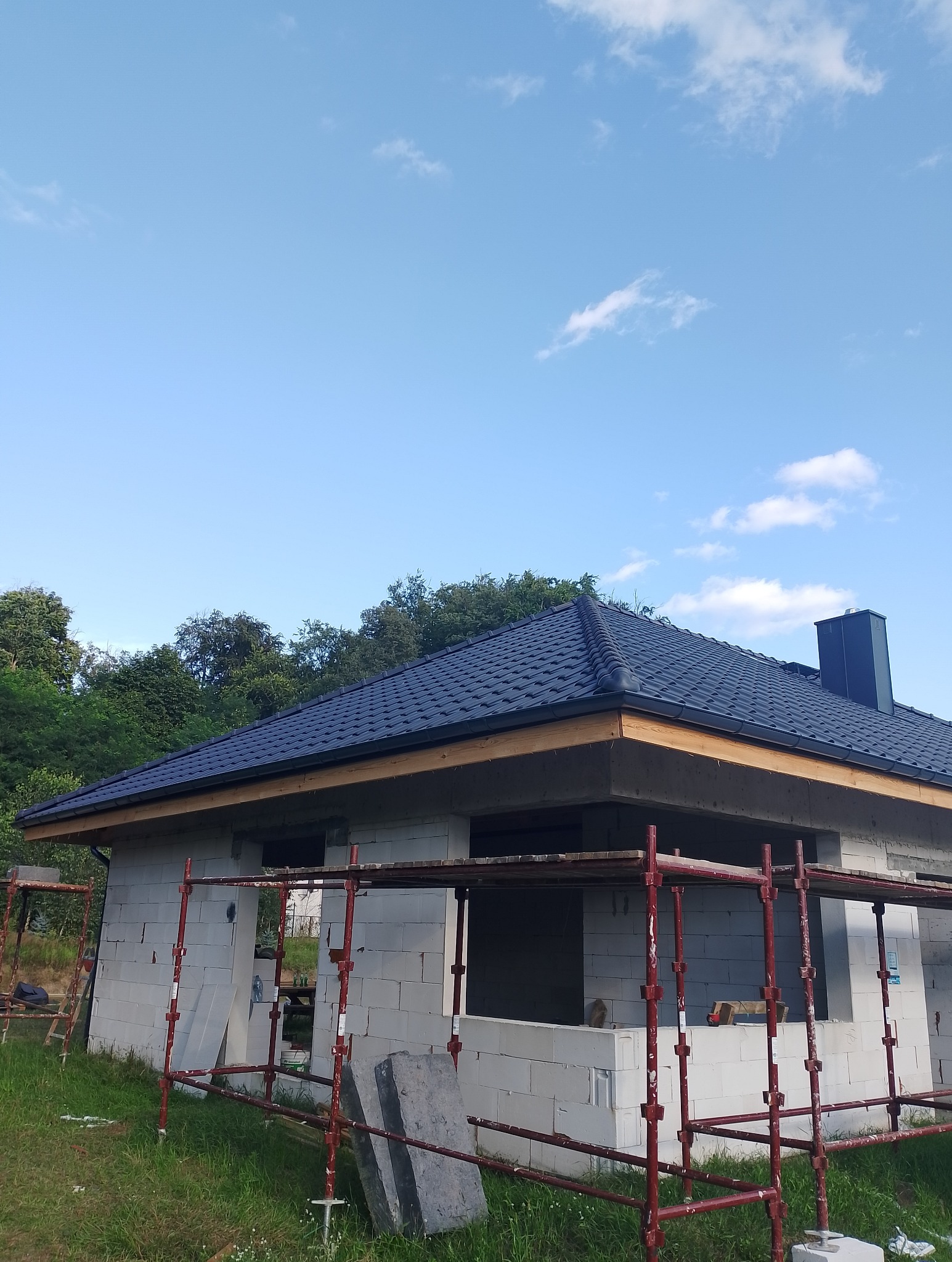 Niedokończony dom z ciemnym dachem z dachówki ceramicznej, otoczony rusztowaniem i zielenią, z widocznym kominem i błękitnym niebem w tle.