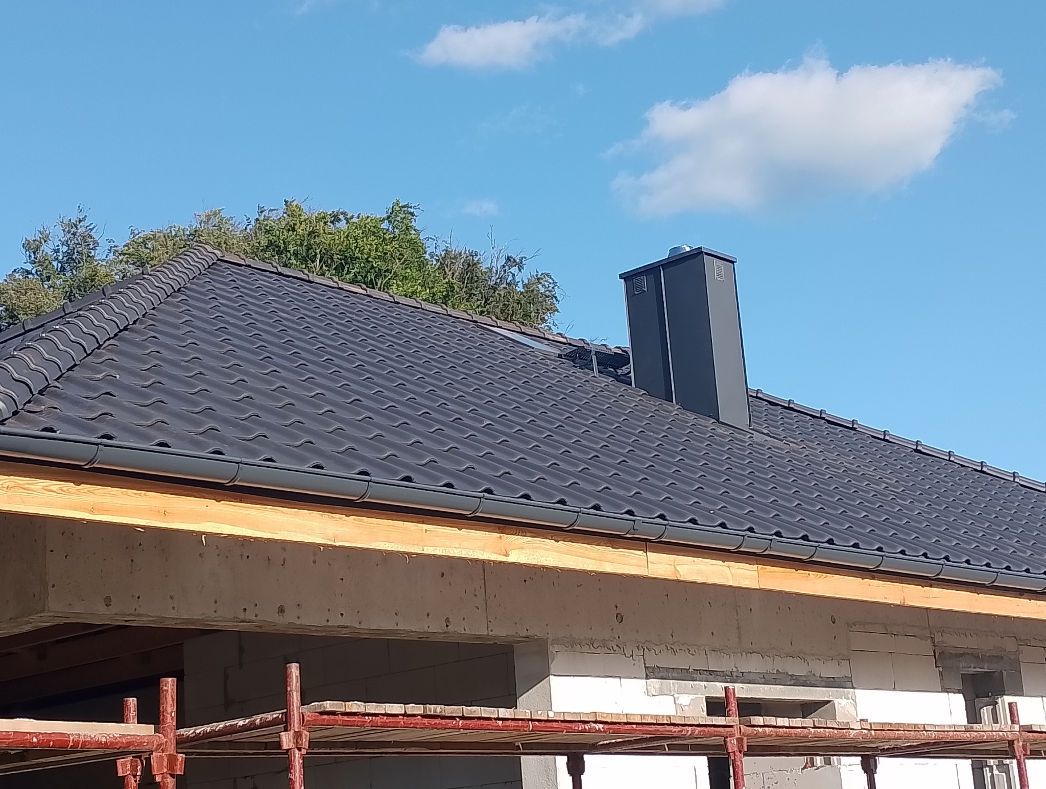 Nowy dach z ciemnej dachówki ceramicznej i szarym kominem na tle błękitnego nieba z chmurami, widoczne rusztowanie i surowy stan budynku.