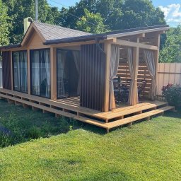 Sviatlana Ausiannikava - Drewniana konstrukcja z zadaszeniem i oknami, częściowo osłonięta pionowymi listwami i zasłonami, umieszczona na podwyższonym tarasie z drewna na trawniku.