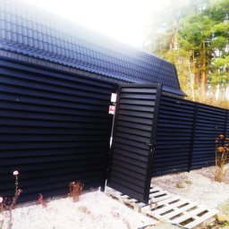 Sviatlana Ausiannikava - Nowoczesne, czarne ogrodzenie panelowe z otwartą furtką, ułożoną na palecie, na tle białego żwiru i domu z ciemnym dachem.