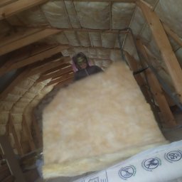 Sviatlana Ausiannikava - Osoba z maską na twarzy instaluje izolację z wełny mineralnej na poddaszu z drewnianą konstrukcją więźby dachowej, widoczna drabina i opakowania izolacji.