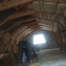 Sviatlana Ausiannikava - Pracownik w trakcie ocieplania poddasza wełną mineralną, widoczne drewniane krokwie, okno i częściowo wykończona ściana z cegły.