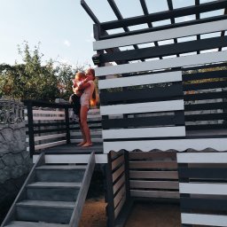 Sviatlana Ausiannikava - Dzieci na drewnianej konstrukcji z elementami pomalowanymi na czarno i biało, przypominającej domek lub plac zabaw, z schodami i ogrodzeniem z imitacji kamienia w tle.