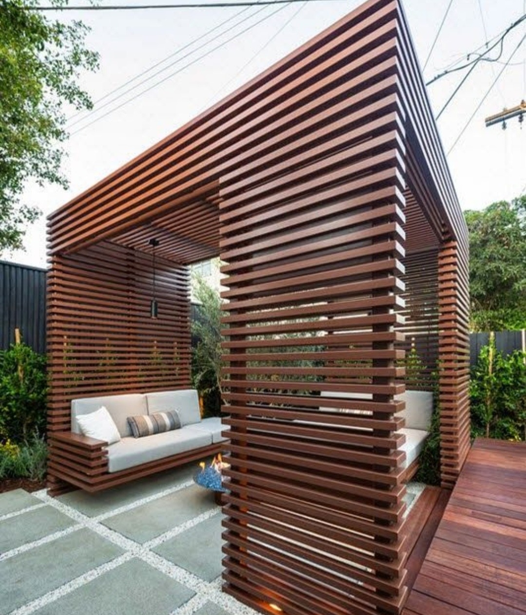 Nowoczesna konstrukcja z drewnianych listew tworząca przestrzeń wypoczynkową z sofą, paleniskiem i drewnianym tarasem, otoczona zielenią.