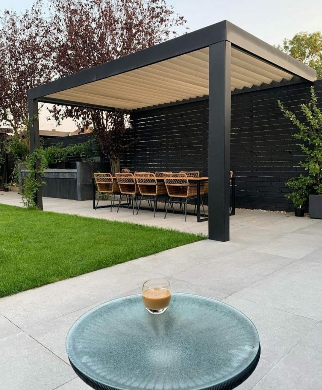 Nowoczesna pergola z czarną ramą i beżowym zadaszeniem lamelowym, widok z perspektywy niskiego stolika z napojem, w tle czarne ogrodzenie i zielony trawnik.