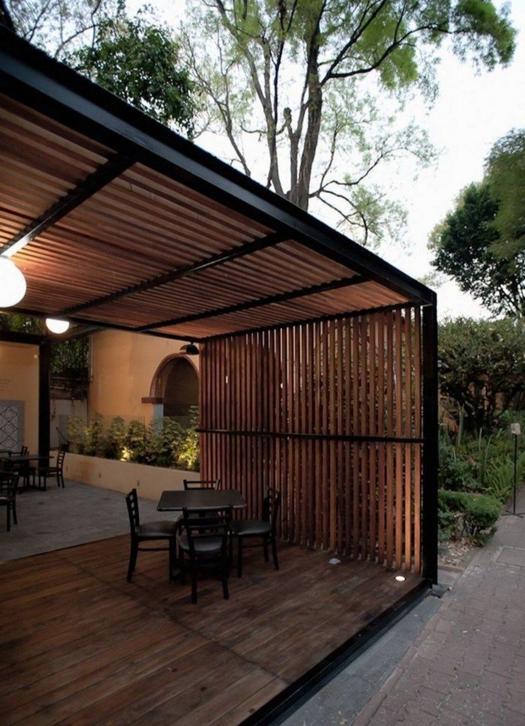Nowoczesna konstrukcja zadaszenia tarasu z drewnianymi listwami i czarną metalową ramą, oświetlona lampami, z drewnianą podłogą i zestawem mebli ogrodowych w tle zieleni.