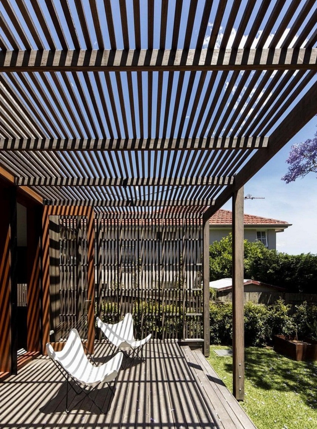 Drewniana pergola z ażurowym dachem rzucająca cienie na taras z desek, na którym stoją dwa białe krzesła typu motyl. W tle widoczny ogród z zielenią i dom z czerwoną dachówką.
