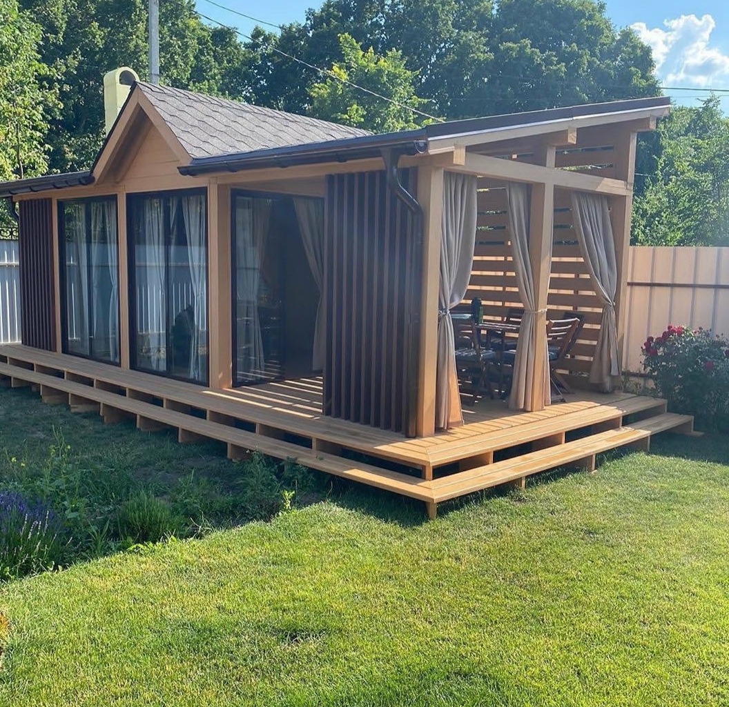 Drewniana konstrukcja z zadaszeniem i oknami, częściowo osłonięta pionowymi listwami i zasłonami, umieszczona na podwyższonym tarasie z drewna na trawniku.
