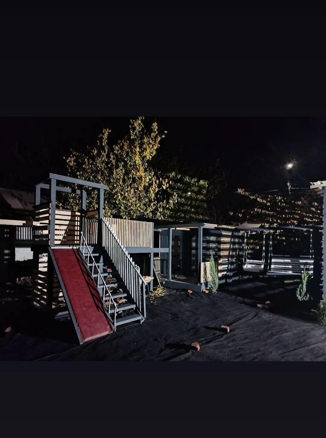 Drewniany plac zabaw z czerwoną zjeżdżalnią i huśtawką, oświetlony nocą, z widocznymi elementami konstrukcyjnymi i drobnymi roślinami.