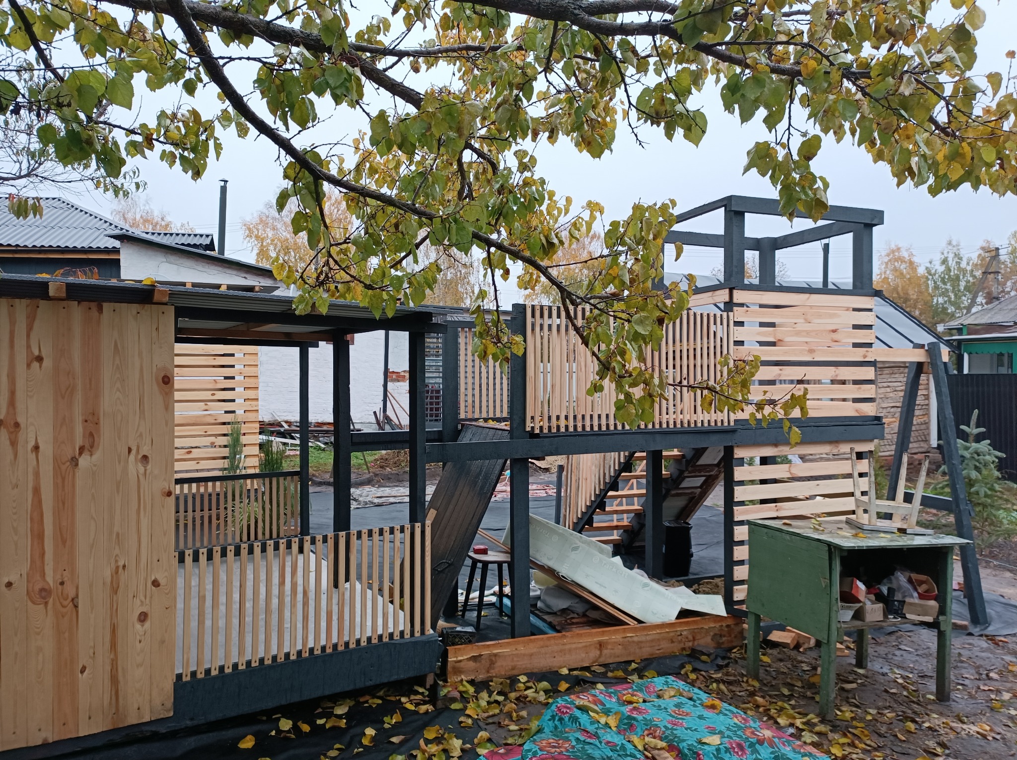 Konstrukcja drewniana w trakcie budowy, dwupoziomowa, z elementami altany i tarasu, widoczne rusztowania, narzędzia i materiały budowlane, jesienne liście na ziemi.
