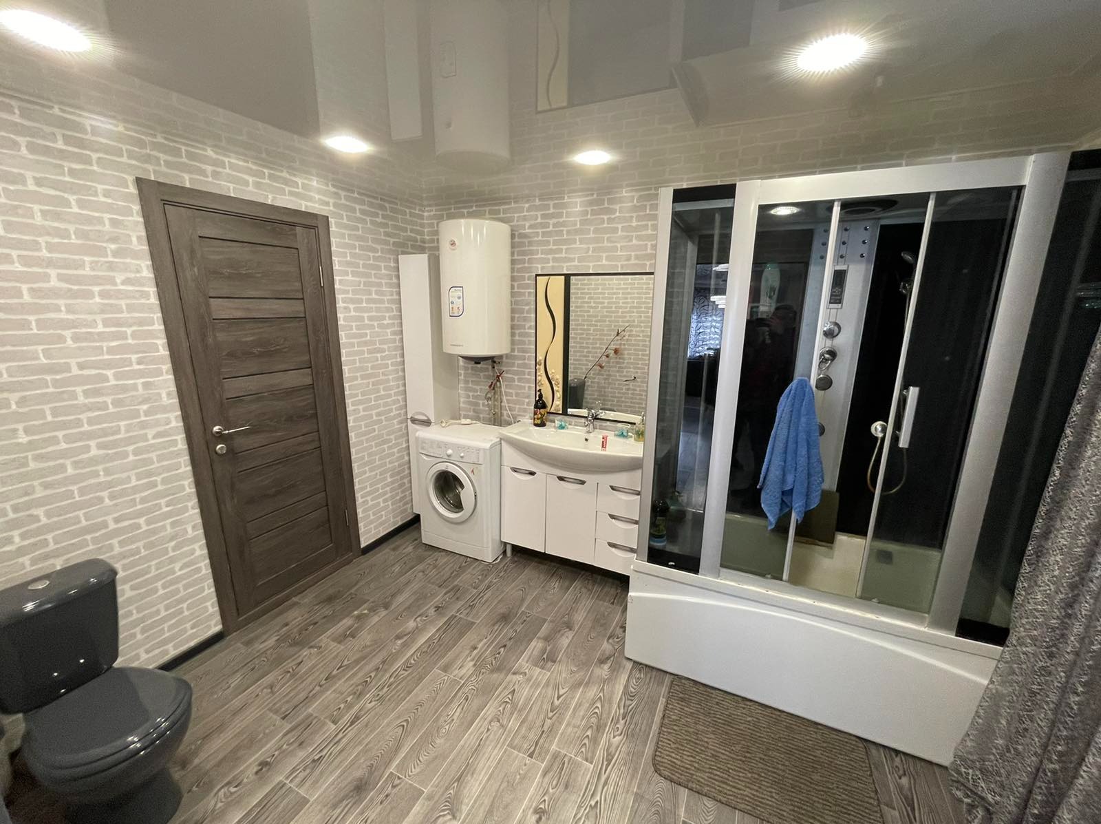Wyremontowana łazienka w szaro-białej tonacji, z kabiną prysznicową z hydromasażem, umywalką z szafką, pralką, bojlerem i toaletą w kolorze grafitowym.