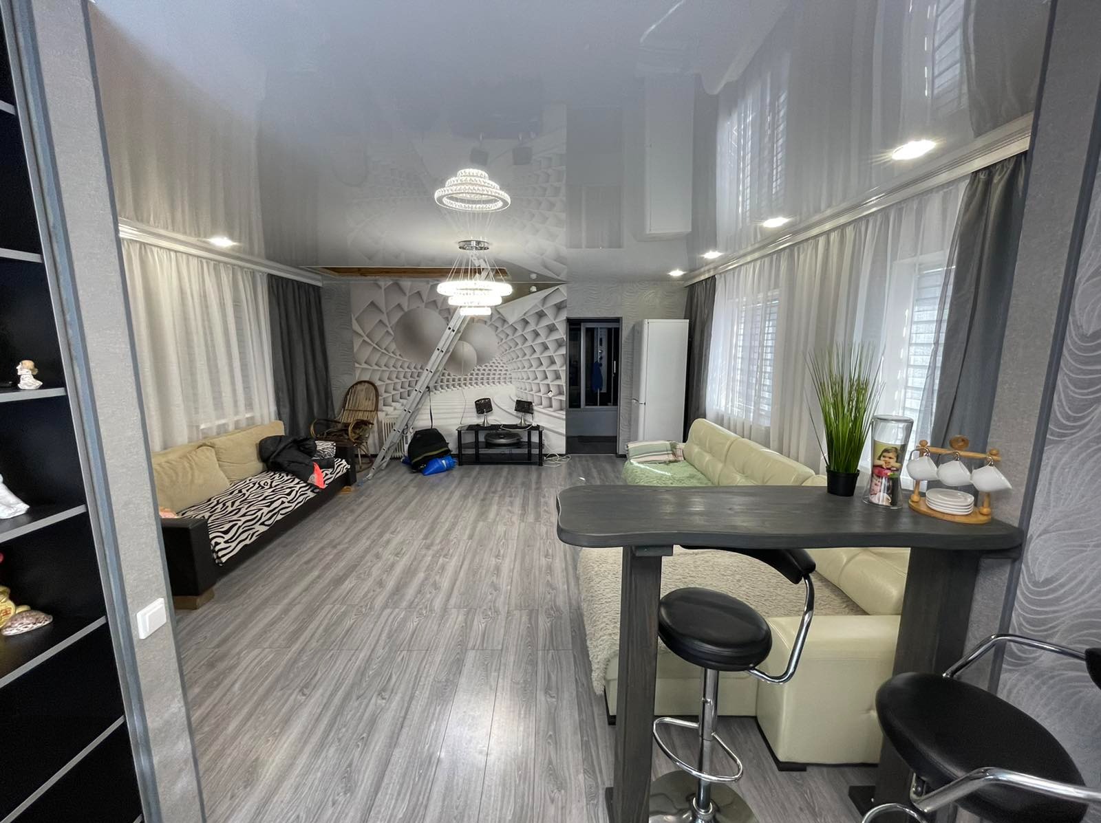 Wnętrze salonu z szarą podłogą imitującą drewno, jasnymi sofami, wysokim połyskliwym sufitem i dekoracyjną ścianą z geometrycznym wzorem, z widoczną drabiną sugerującą prace wykończeniowe.