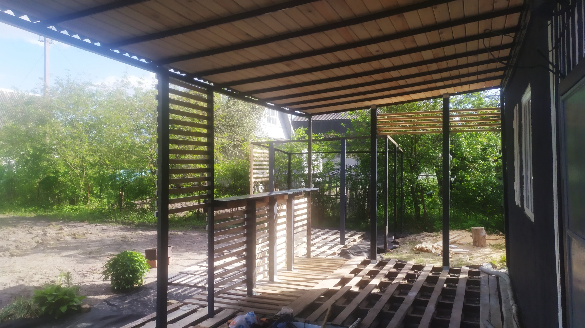 Konstrukcja zadaszenia z drewnianymi elementami i metalowymi słupami w trakcie montażu, widoczne deski podłogowe i tło z zielenią.