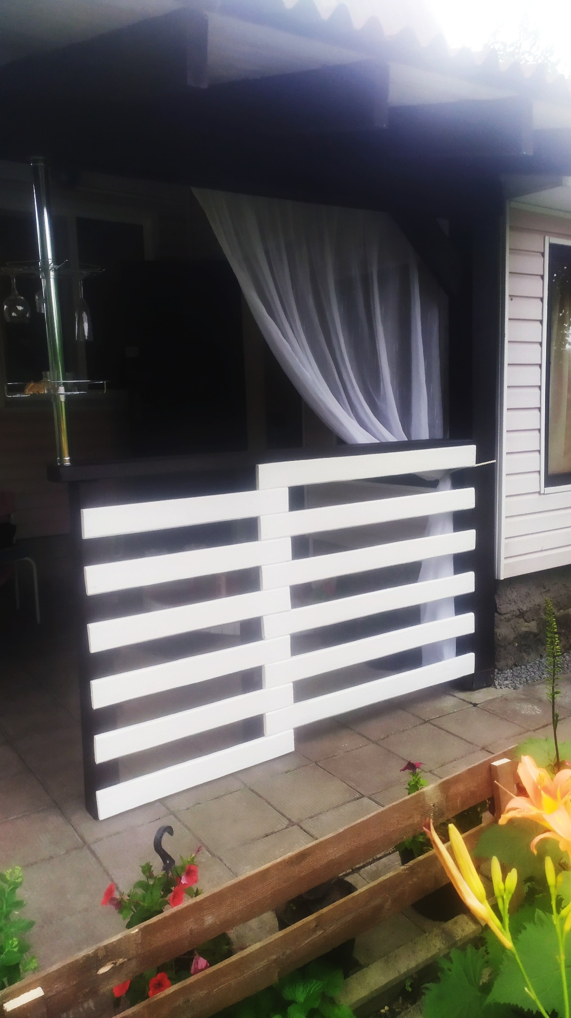 Zadaszony taras z czarną konstrukcją i białymi, poziomymi panelami, zasłonięty delikatną, białą firanką, widok z zewnątrz przez niskie, drewniane ogrodzenie z kwiatami.