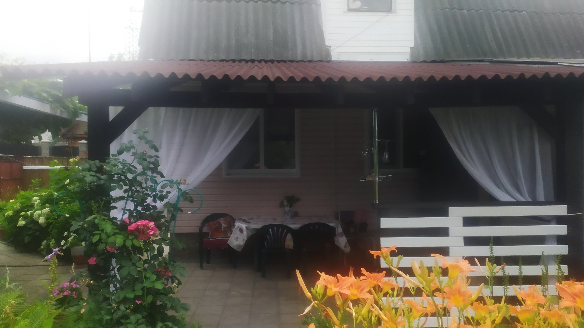 Zadaszony taras z ciemnego drewna, zasłonięty białymi firanami, z widocznym stołem i krzesłami, otoczony kwitnącymi kwiatami w odcieniach różu i pomarańczy.