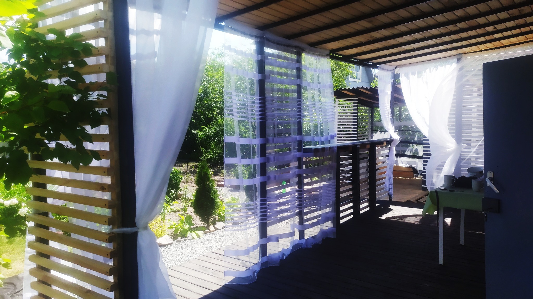 Drewniana pergola z białymi zasłonami, tworząca zacienioną przestrzeń na zewnątrz z widokiem na ogród.