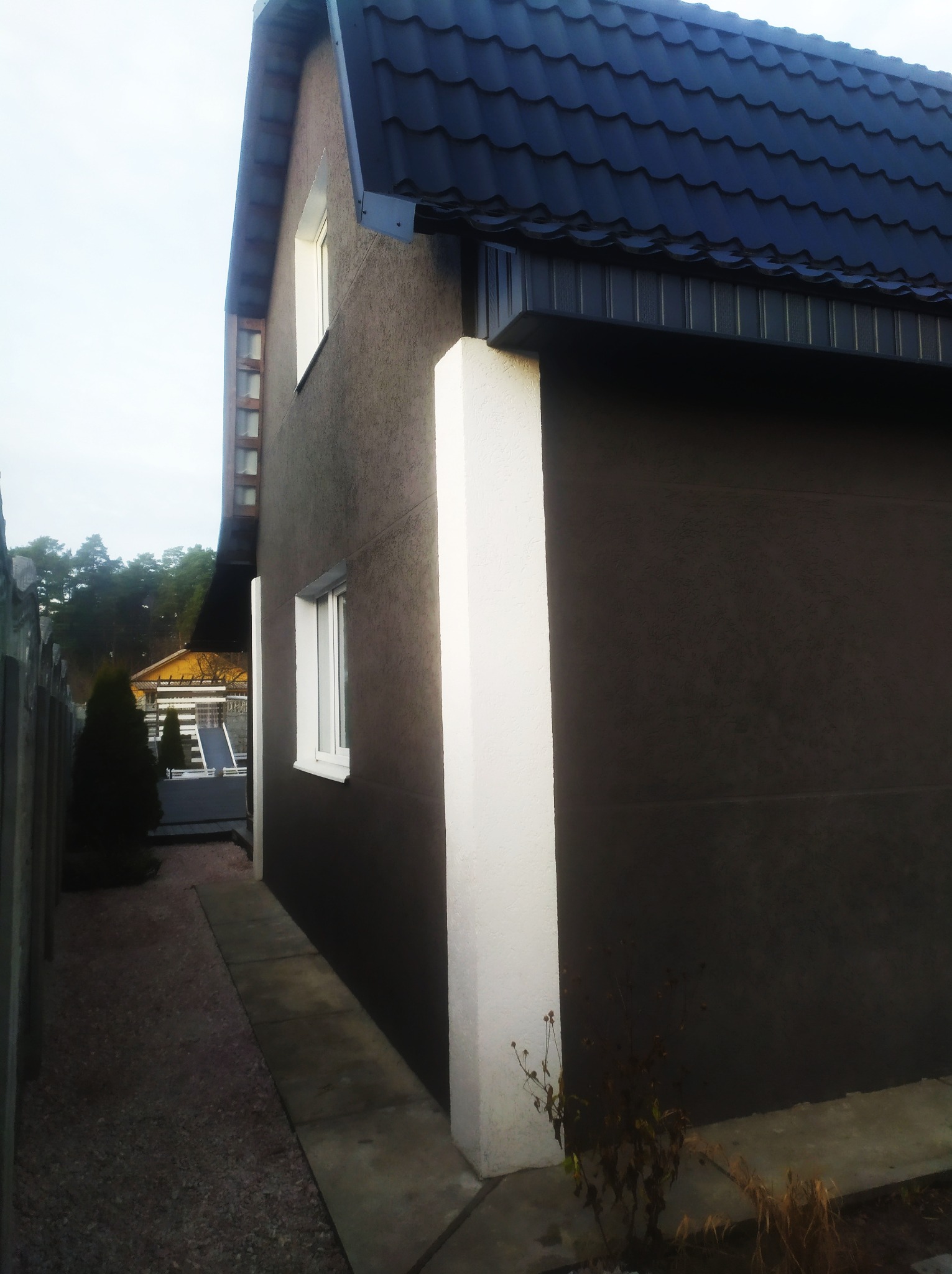 Boczny widok domu z ciemnobrązową elewacją i białymi narożnikami, czarny dach z metalowej blachy, widoczny chodnik i fragment ogrodu z tujami w tle.